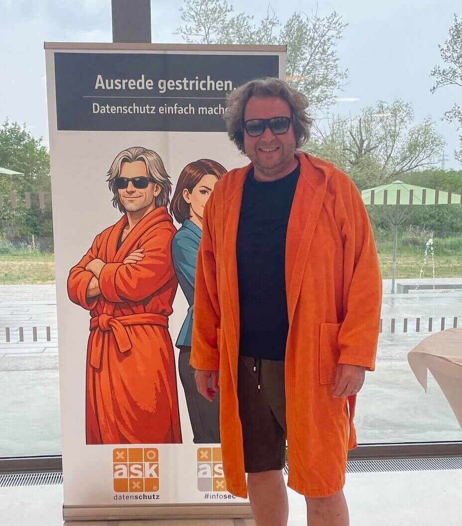 Sascha Kuhrau, verkleidet als der Datenschutz Dude im orangefarbenen Bademantel, lächelnd vor einem Banner mit der Aufschrift "Ausrede gestrichen. Datenschutz einfach machen."